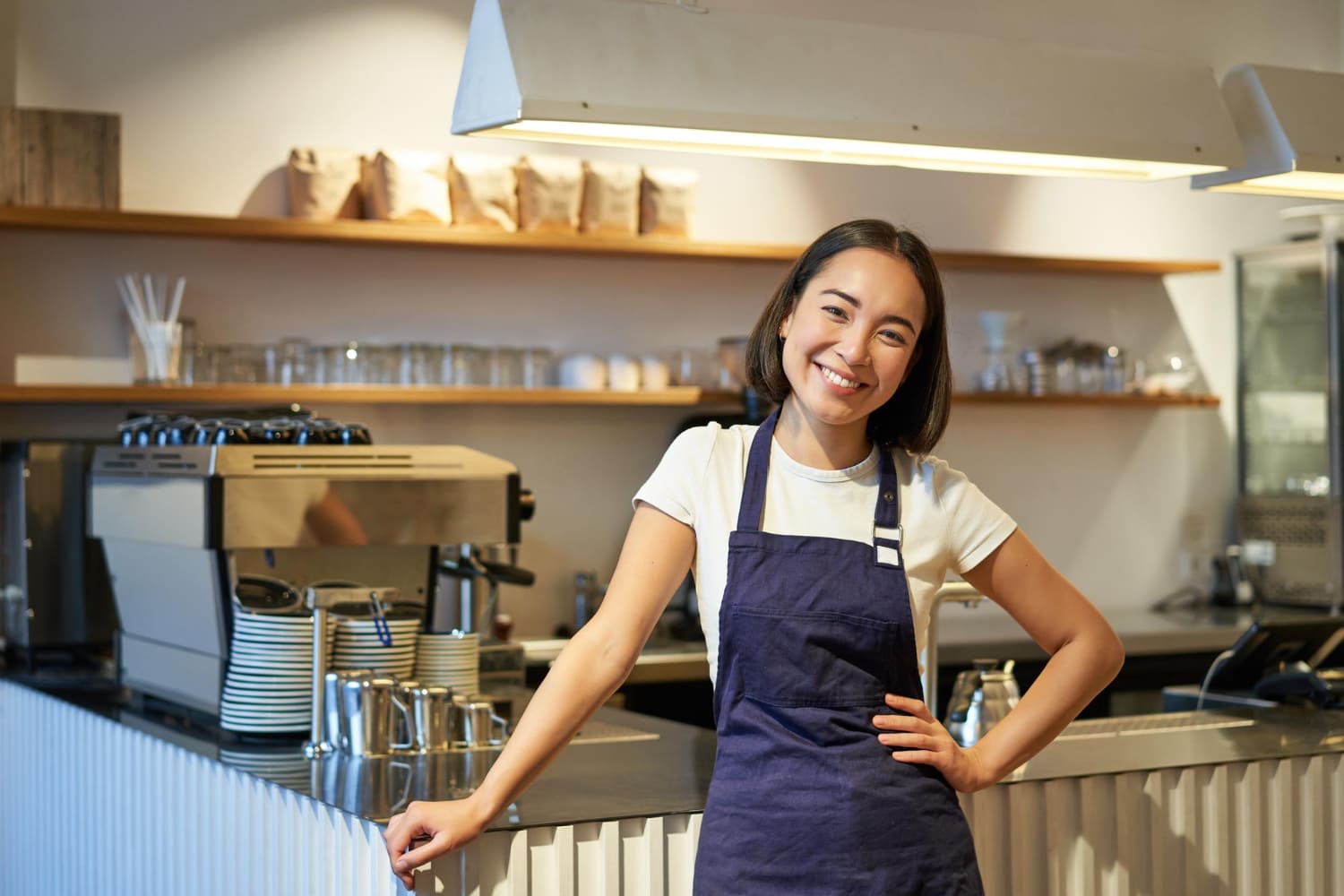 joven-barista-sonriente-delantal-trabajando-cafe-parado-cerca-mostrador-maquina-cafe-mirando